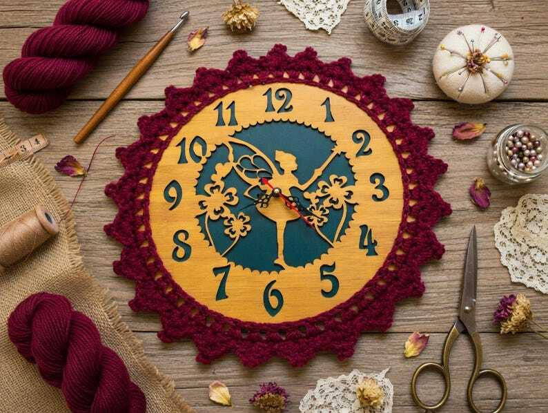 Wooden wall clock with fairy silhouette and burgundy crocheted edge, surrounded by yarn, a crochet hook, scissors, measuring tape, pins and other sewing supplies on a wooden table.