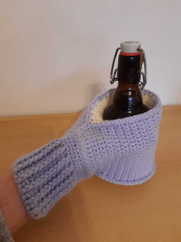 Hellviolette gehäkelte Flaschenhülle mit integrierter Armstulpe umschließt eine braune Bügelverschlussflasche.