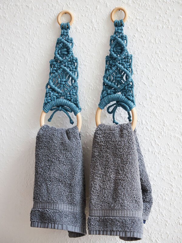 Zwei petrolfarbene Makramee-Handtuchhalter mit Holzringen und grauen Handtüchern vor weißer Wand.