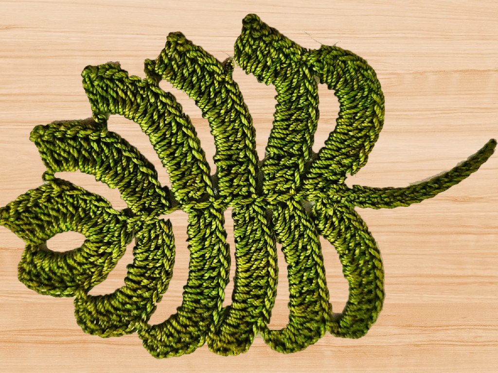 Green crocheted leaf-shaped coaster with textured stitches on a light wood surface.