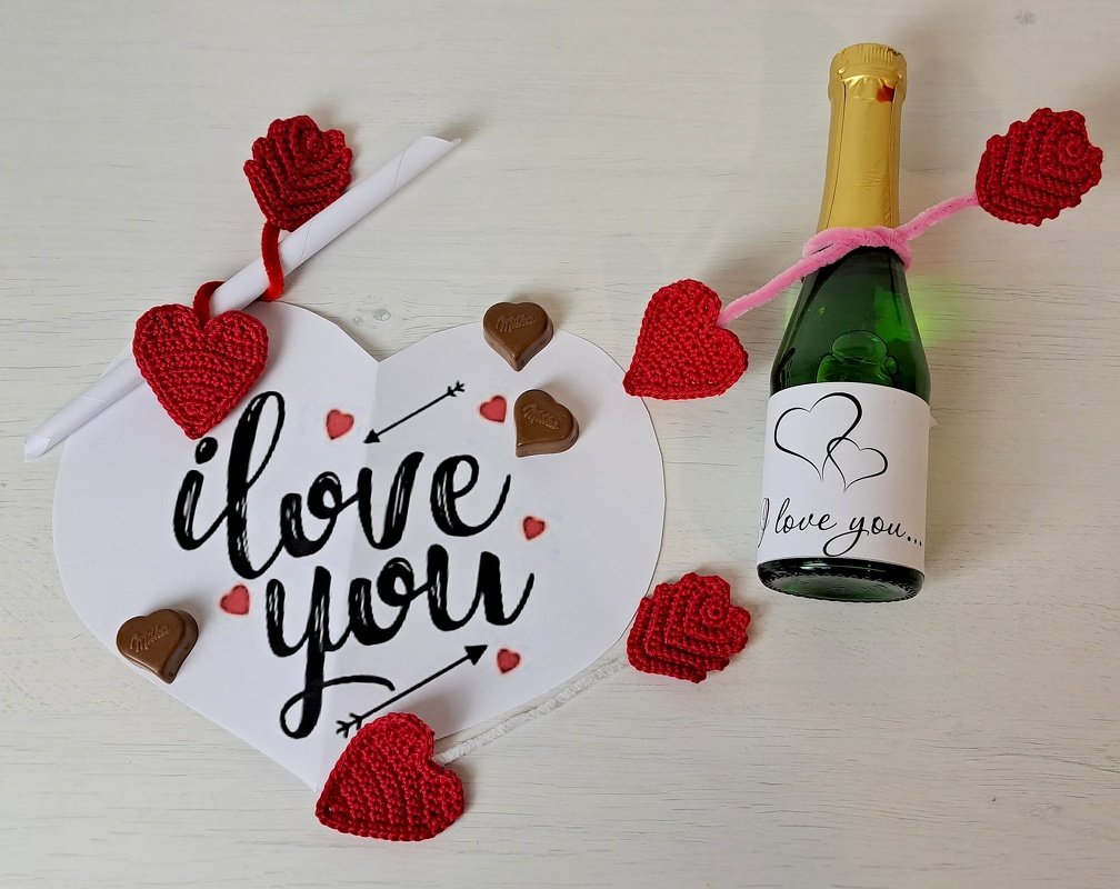 Gehäkelte rote Herzen und Rosen neben einer Herzkarte mit 'I love you', Schokoladenherzen und einer dekorierten Flasche auf hellem Untergrund.
