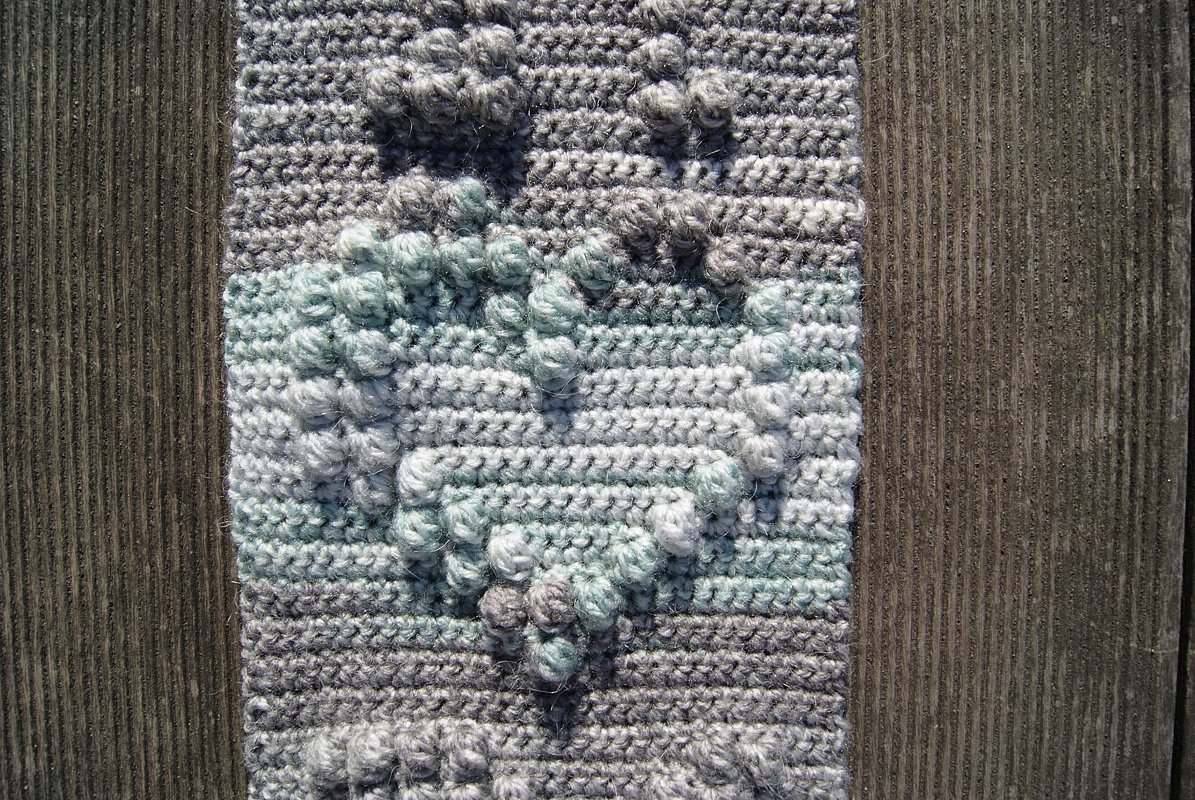 Nahaufnahme eines gehäkelten Stücks mit erhabenen Bobbelstichen in Grau, Weiß und Mint auf Holz.