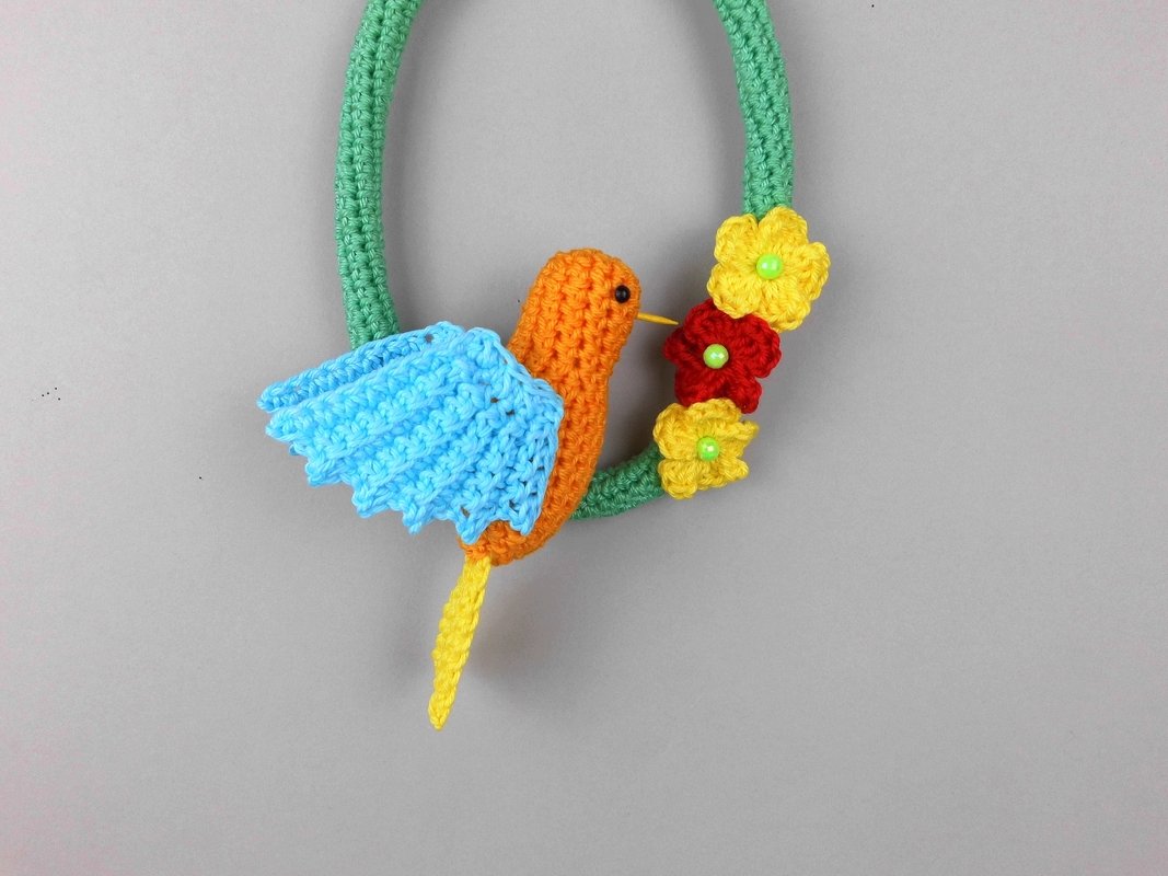 Gehäkelter Kolibri mit orangefarbenem Körper und hellblauen Flügeln an einem grünen Häkelkranz mit gelben und roten Blumen und grünen Perlen.