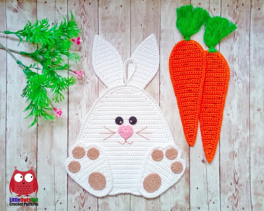 White crocheted bunny wall hanging with stitched paw pads and two orange crocheted carrots on a light wooden background