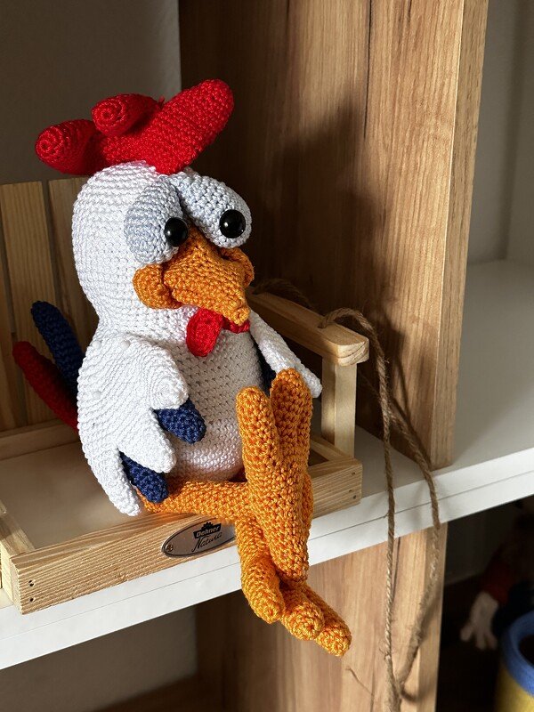 Gehäkelter Amigurumi-Hahn mit rotem Kamm auf Holzregal