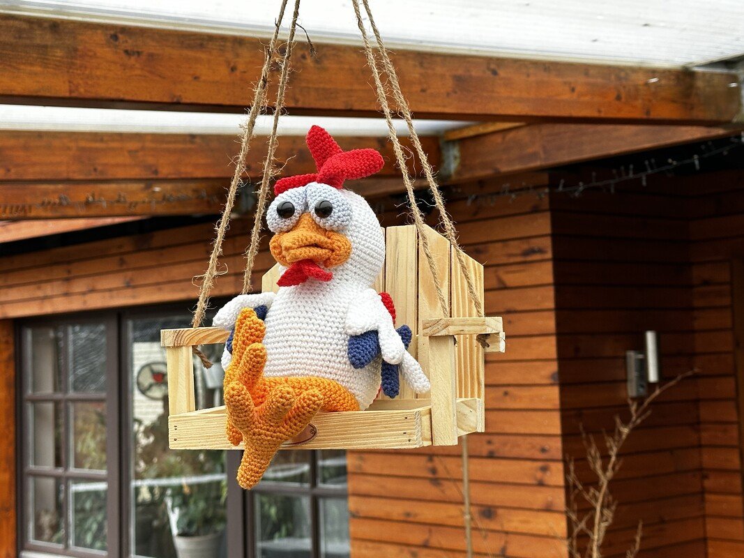 Gehäkeltes weiß-oranges Huhn mit rotem Kamm auf hölzerner Hängeschaukel