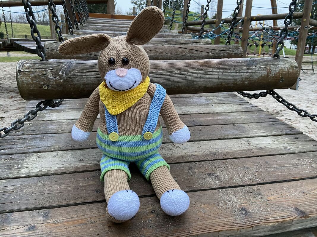 Gestrickter Amigurumi-Hase mit gelbem Halstuch und gestreifter Latzhose, sitzend auf einem hölzernen Spielplatzsteg