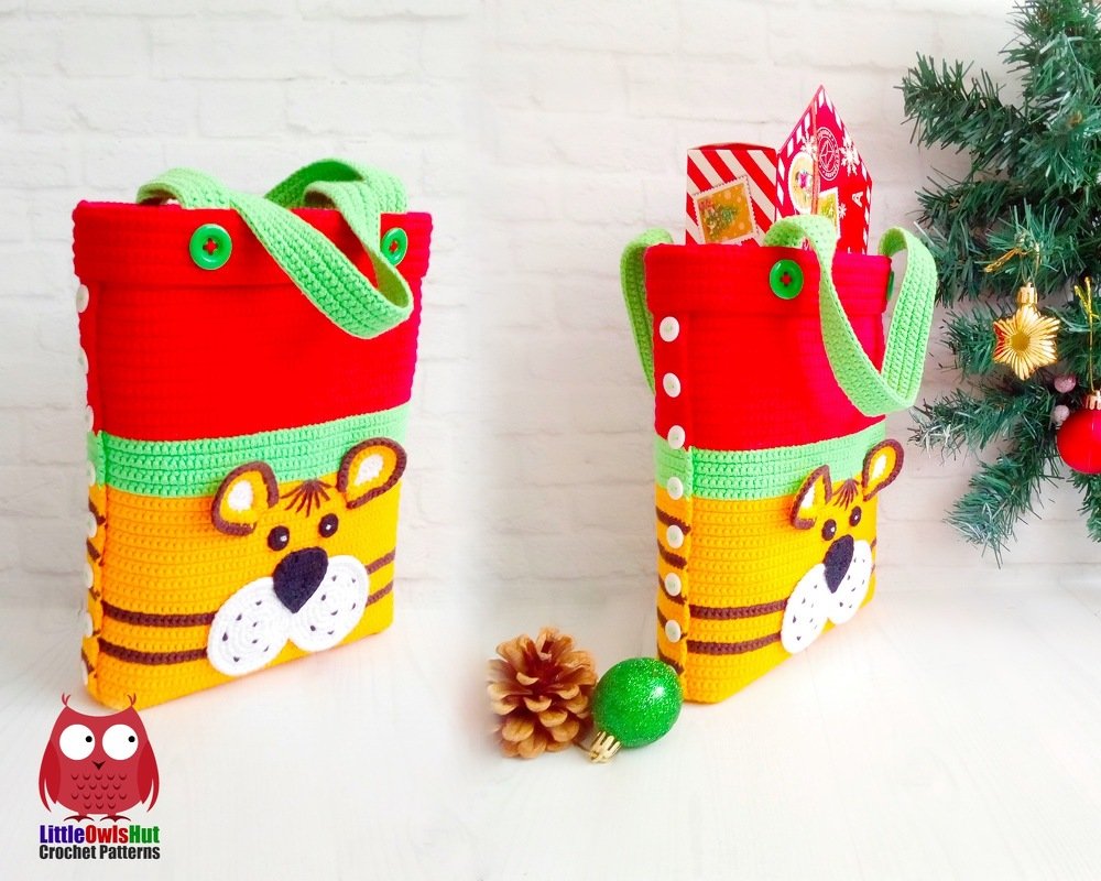 Bright red, yellow and green crochet tote with a tiger face appliqué, green handles and white side buttons, styled beside a small decorated tree and pine cone.