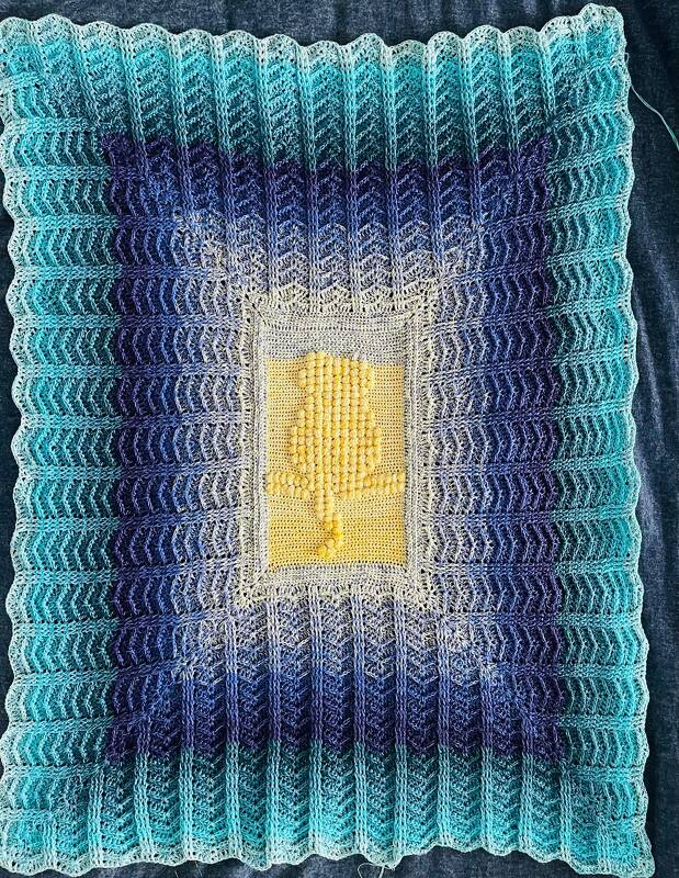 Gehäkelte Decke mit türkis-blauem Farbverlauf und gelbem, reliefartigen Bobble-Mittelteil