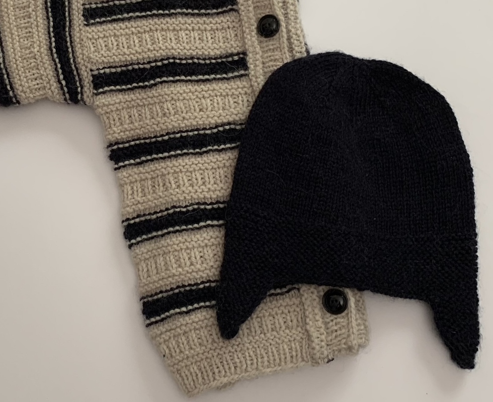 Gestrickte beigefarbene Babyjacke mit dunklen Streifen neben einer dunklen Babymütze