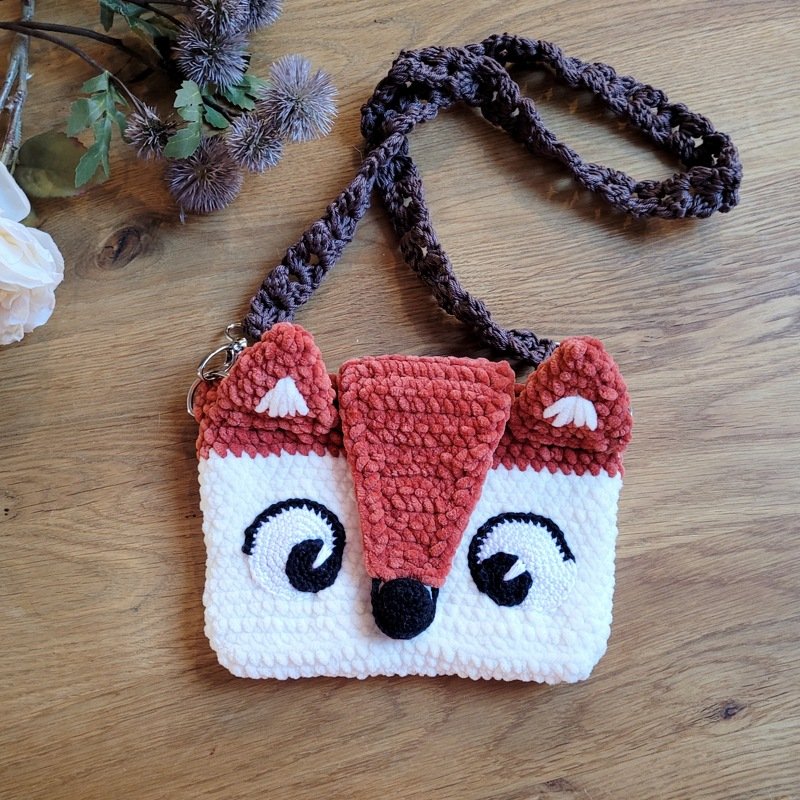 Flauschige weiß-rostbraune Häkeltasche mit Fuchsgesicht und dunkelbraunem Trageriemen auf einem Holztisch.