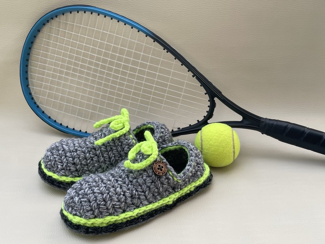 Gehäkelte Babyschuhe im Sneaker‑Stil, grau mit neongrünen Akzenten, neben Tennisschläger und Tennisball.