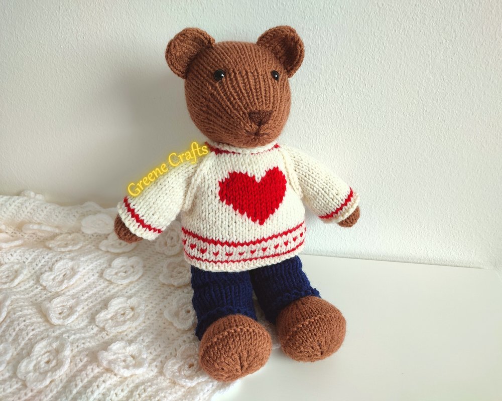 Brown knitted teddy bear wearing a cream sweater with a red heart and blue pants, seated on a white surface.