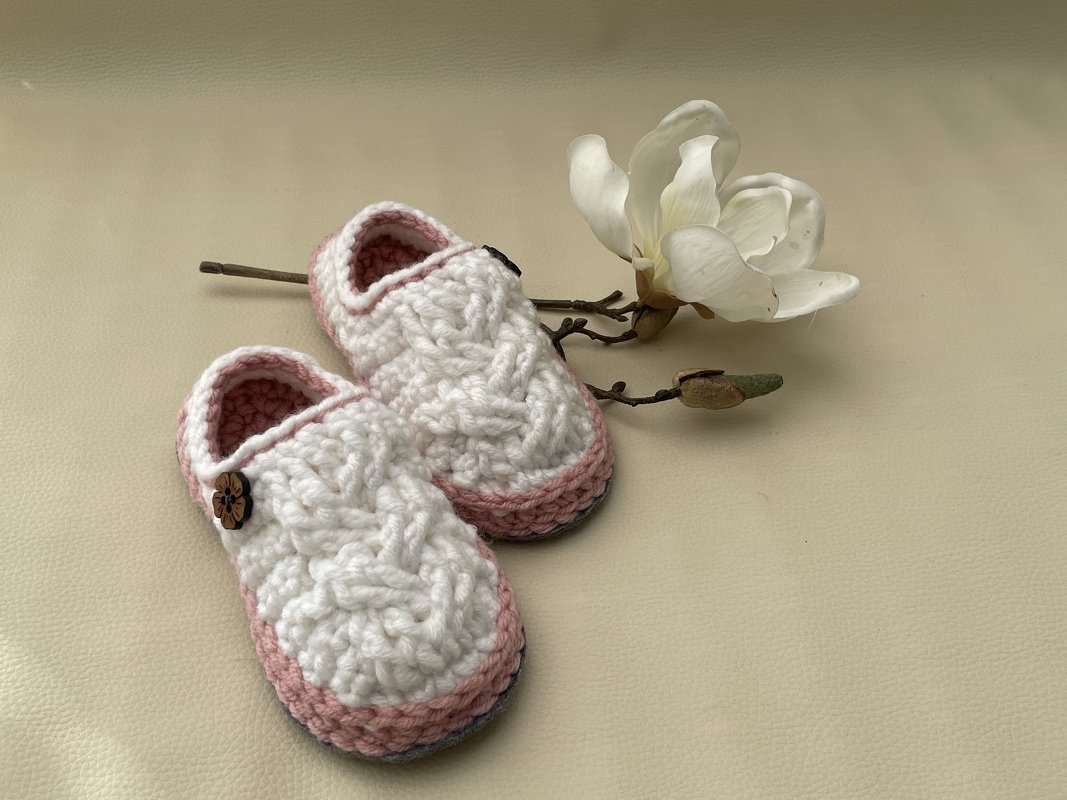 Gehäkelte Babyschuhe in Weiß und Rosa mit Zopfmuster und Holzblumenknopf neben einer Magnolienblüte