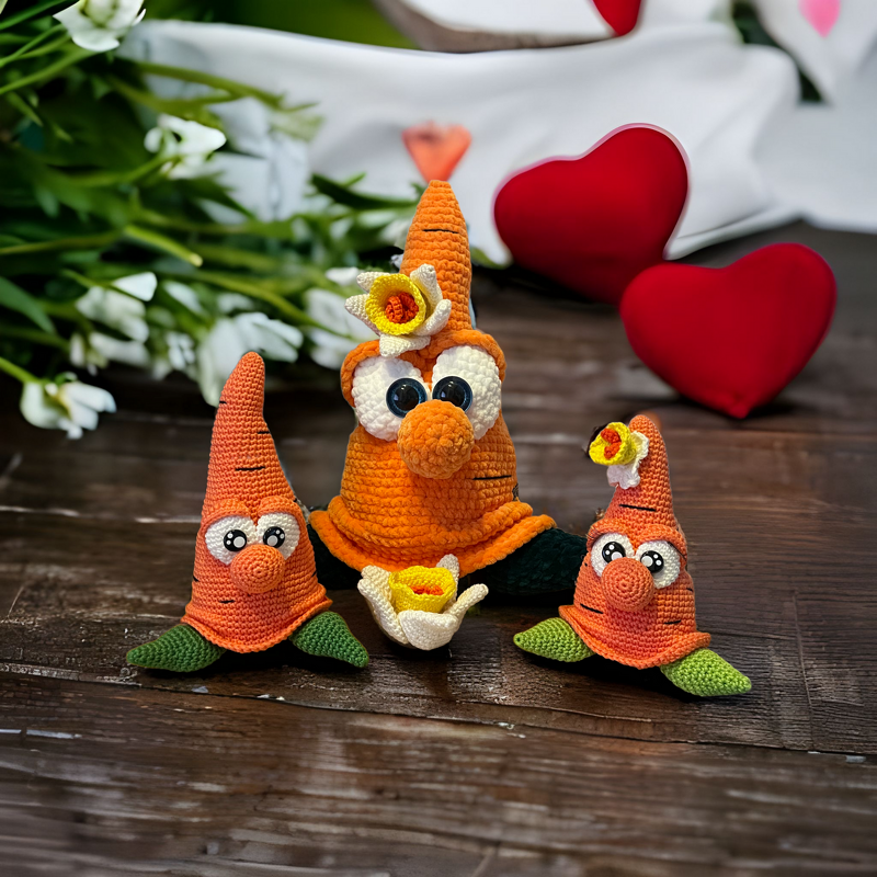 Drei orange gehäkelte Karotten-Amigurumi mit Blüten, grünen Blattfüßen und großen Augen auf einem Holztisch; im Hintergrund rote Herzdekoration