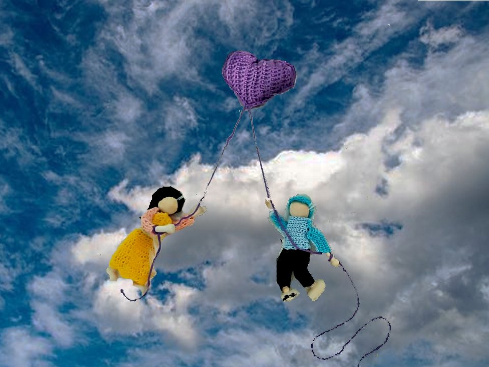 Zwei gehäkelte Puppen halten ein lila Herz und schweben vor bewölktem Himmel.