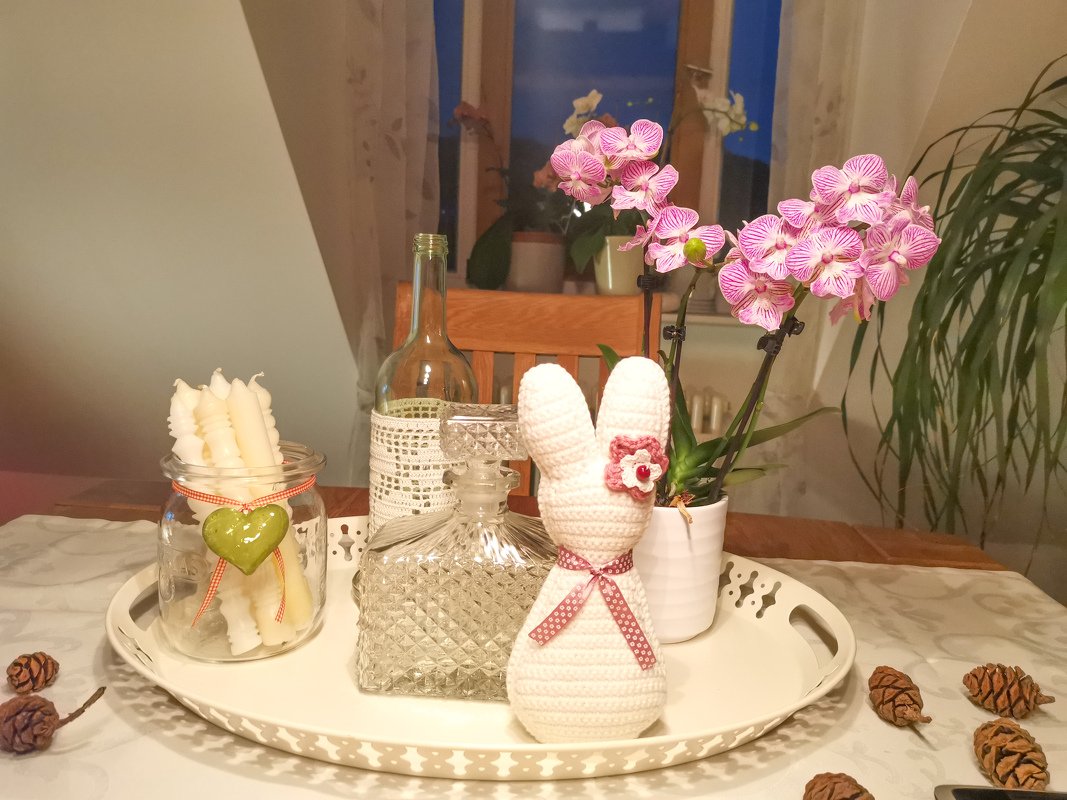 Frühlingshafte Tischdekoration: gehäkelter weißer Hase, Kerzen im Glas, rosa Orchidee und Tannenzapfen auf weißem Tablett.
