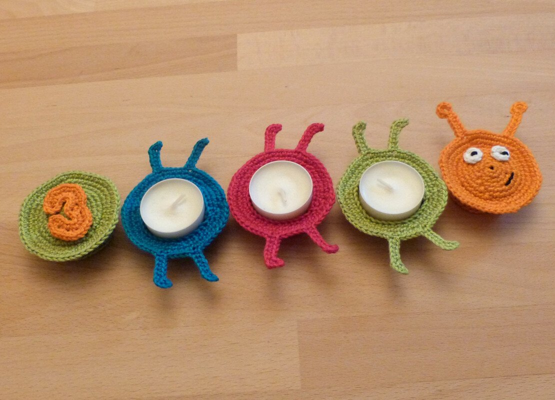 Vier bunte handgehäkelte Monster-Teelichthalter, drei mit Teelichtern, plus gehäkelte Zahl 3 auf hellem Holzuntergrund