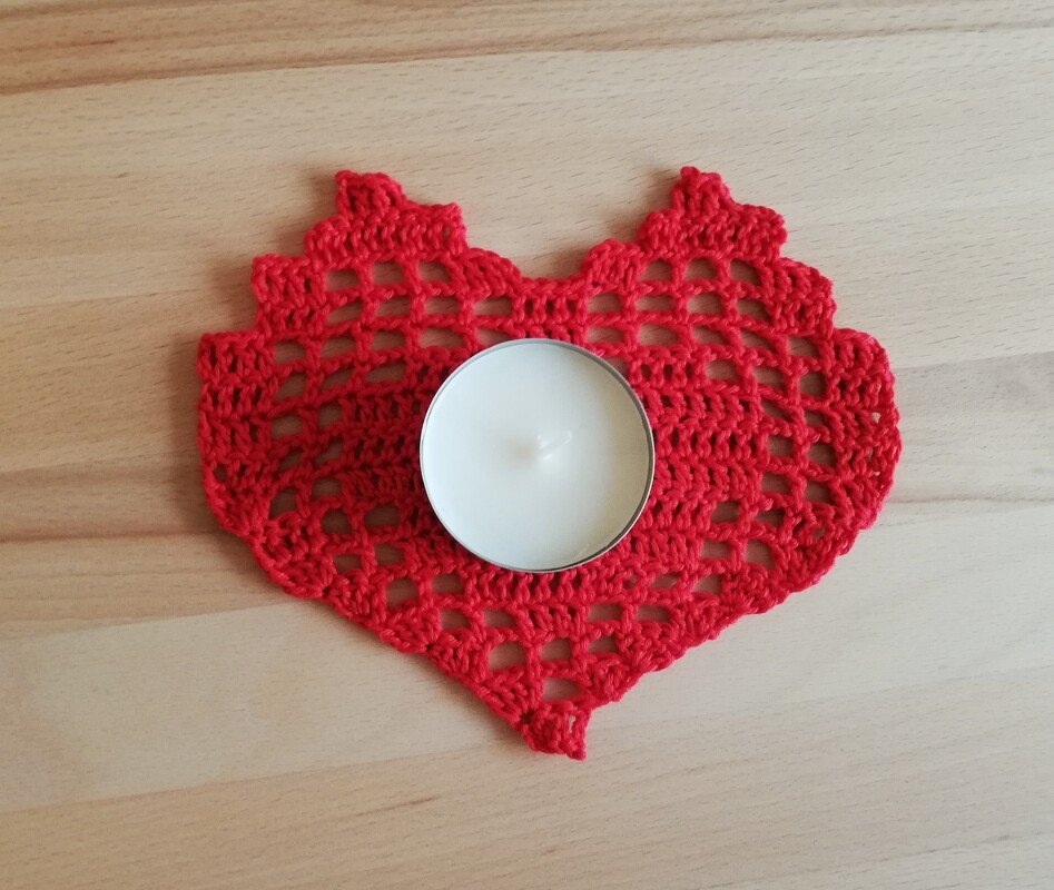 Rotes gehäkeltes Herz-Deckchen mit einem weißen Teelicht in der Mitte auf einer Holzoberfläche.