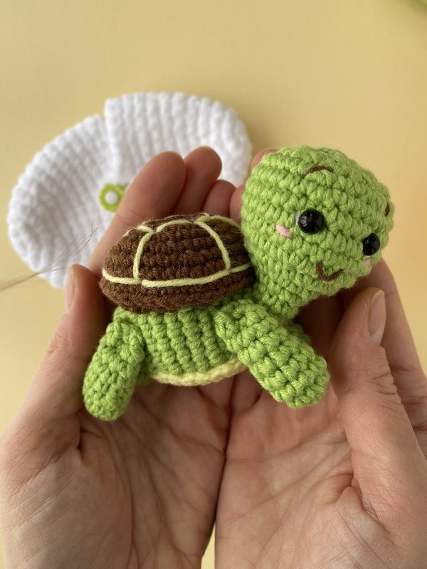 Small green and brown crocheted amigurumi turtle with black eyes and pink cheeks held in hands.