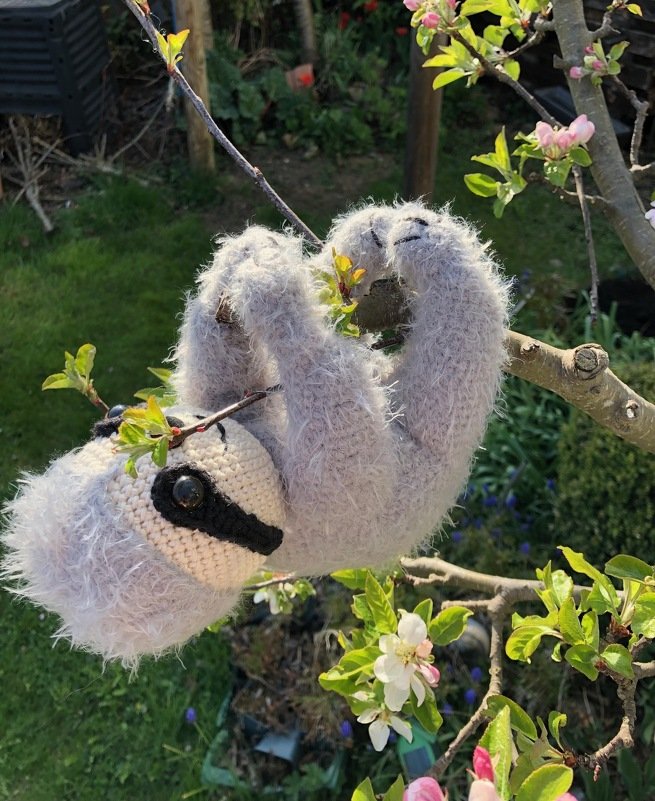 Gehäkeltes flauschiges Faultier-Amigurumi hängt an einem Zweig mit schwarzer Augenmaske