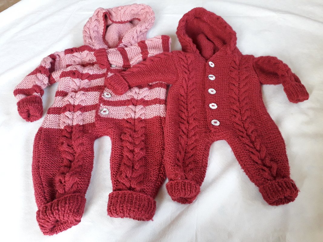 Zwei gestrickte Baby-Overalls in Rot und Rot-Rosa gestreift, beide mit Kapuze, Zopfmuster und Knopfleiste.