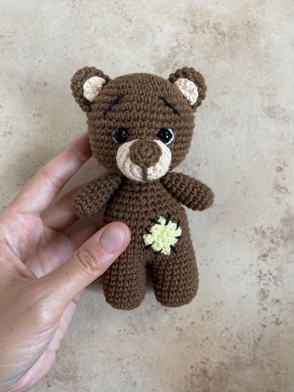 Close-up of a small brown crocheted amigurumi bear with beige muzzle and yellow belly patch held in a hand