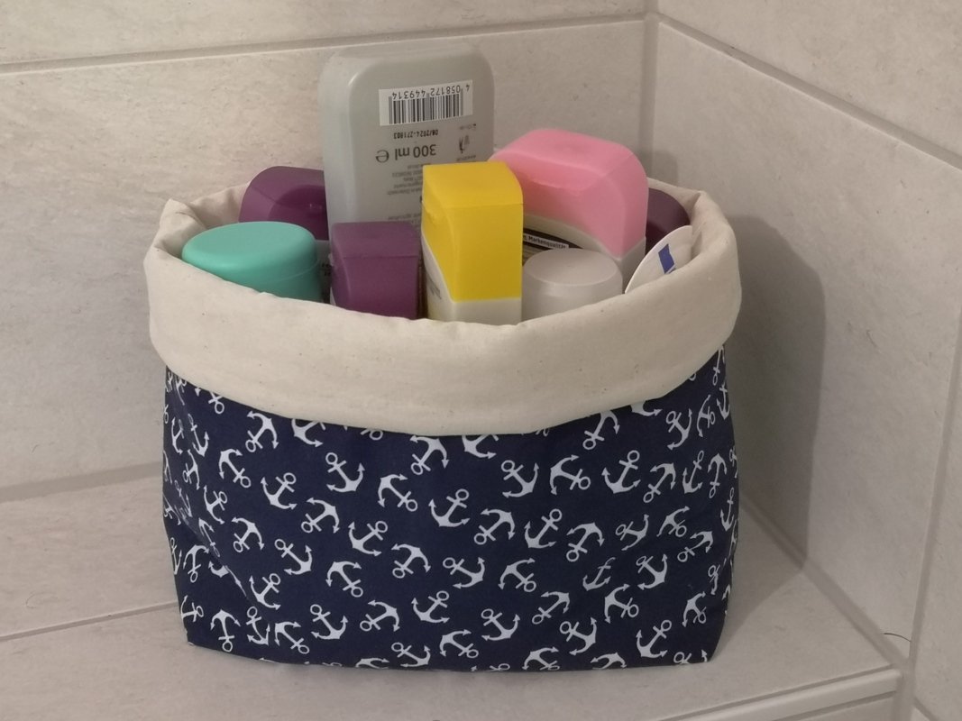 Dunkelblauer Stoffkorb mit weißen Ankern und kremfarbenem Rand, gefüllt mit Hygieneartikeln auf gefliester Ablage