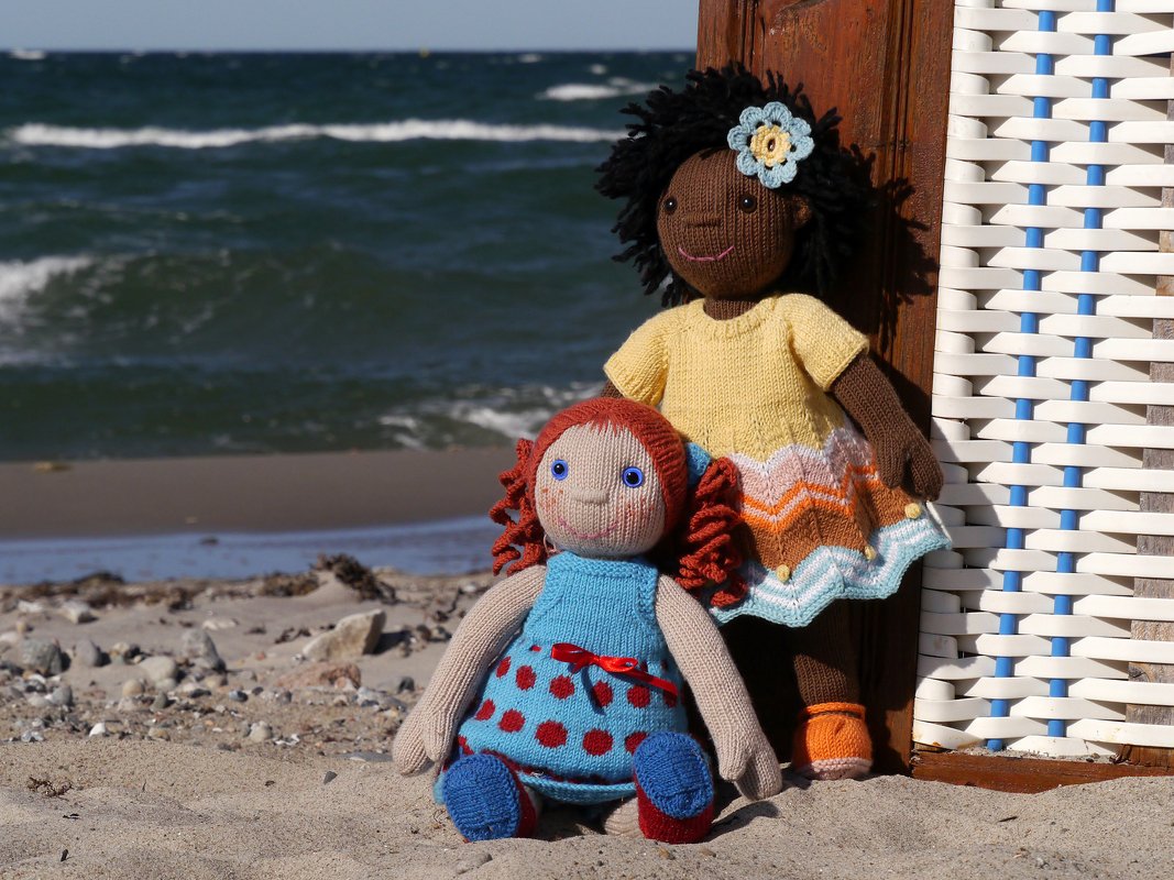 Two knitted dolls on sand beside a white beach chair: red-haired doll in a blue polka-dot dress and a dark-skinned doll in a yellow top with a chevron skirt.