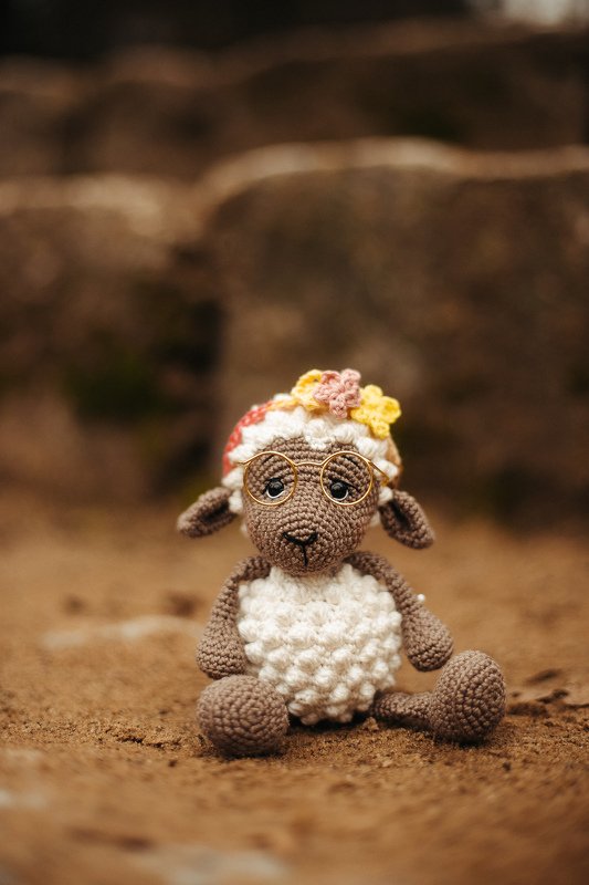 Gehäkeltes Schaf-Amigurumi mit runder Brille und Blumenmütze auf sandigem Untergrund.