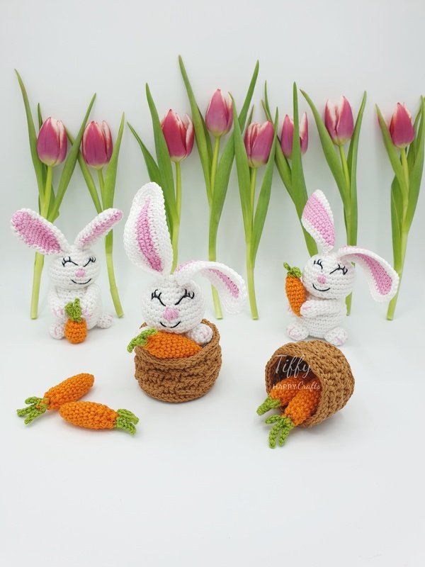 Three white crocheted bunnies with pink ears, several orange crocheted carrots, and pink tulips on a white background.