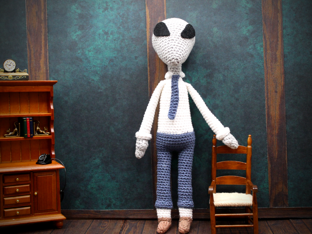 Gehäkelter Alien-Amigurumi in Hemd und Krawatte neben Miniaturmöbeln
