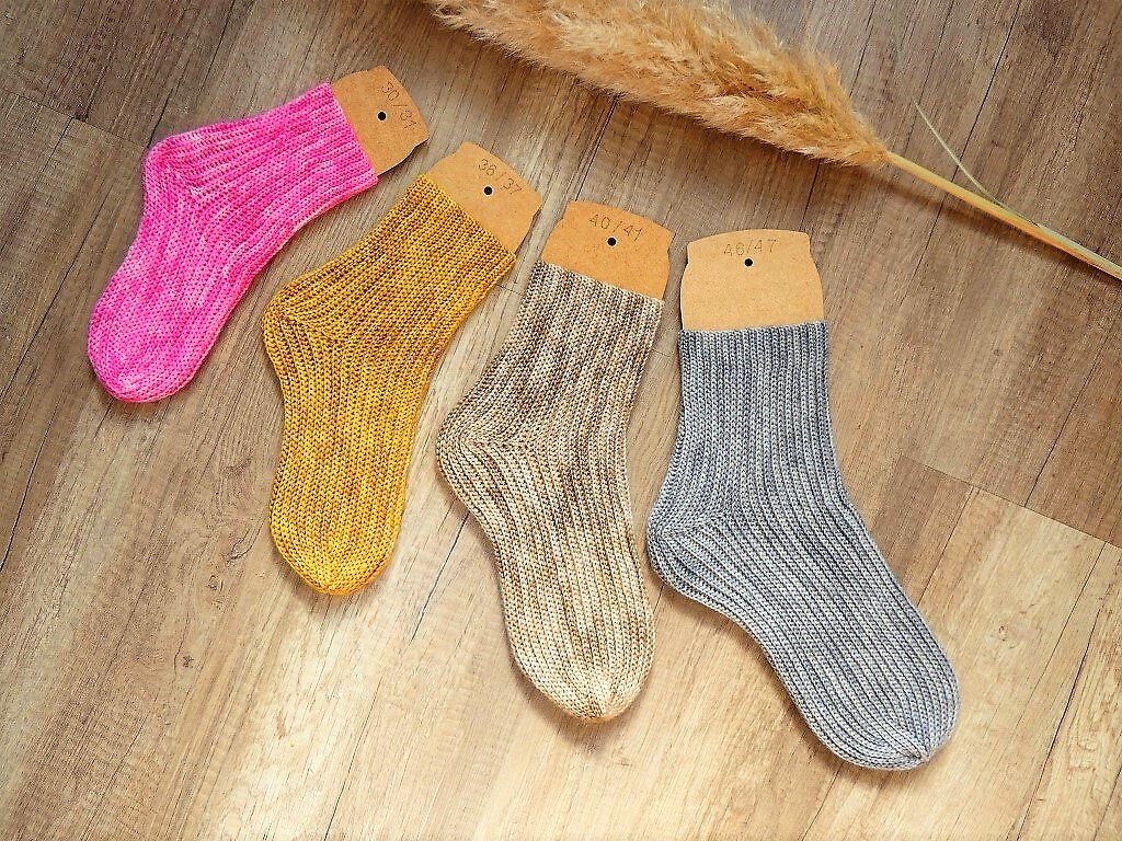 Vier handgehäkelte Rippensocken in Pink, Gelb, Beige und Grau auf Holzboden mit Karton-Einlegern.