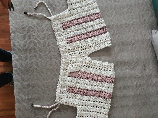 Weißer gehäkelter Bolero mit rosafarbenen Streifen, flach auf einer grauen Decke liegend.