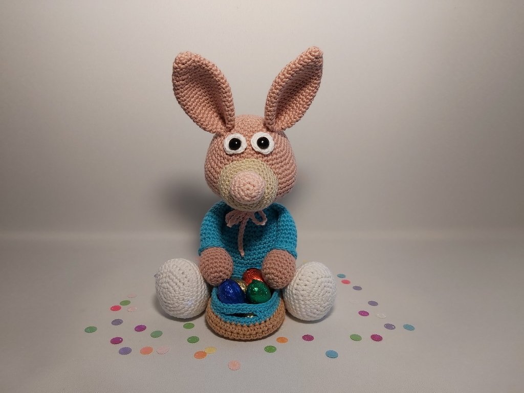 Sitzender Amigurumi-Osterhase mit blauem Pullover und Häkelkörbchen mit bunten Eiern.