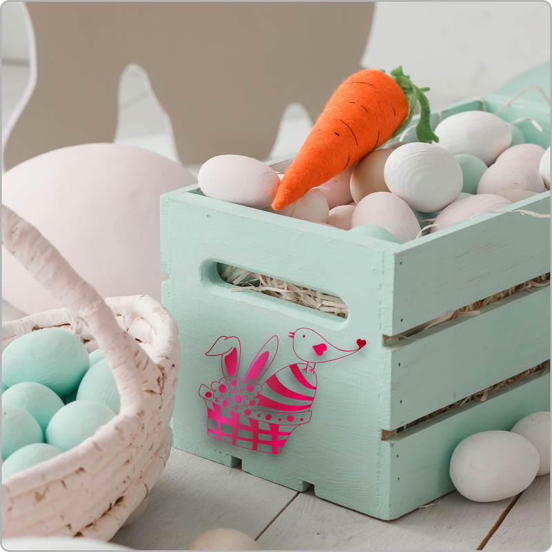 Mintgrüne Holzkiste mit pinkem Hasen- und Vogel-Plottmotiv, gefüllt mit pastellfarbenen Eiern und einer Filzkarotte