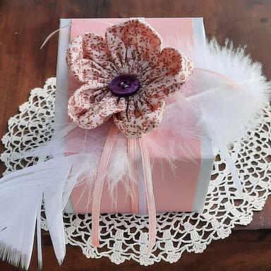 Detailaufnahme: Stoffblume mit lila Knopf, Satinbändern und weißen Federn auf rosa Geschenkbox