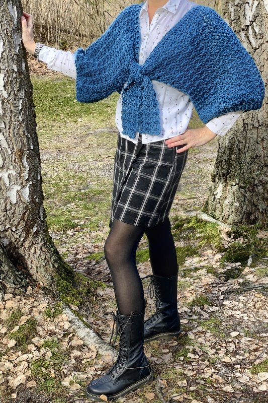 Person trägt gehäkelten blauen Bolero mit vorderer Knotung, kombiniert mit weißem Hemd und kariertem Rock, stehend zwischen Bäumen.