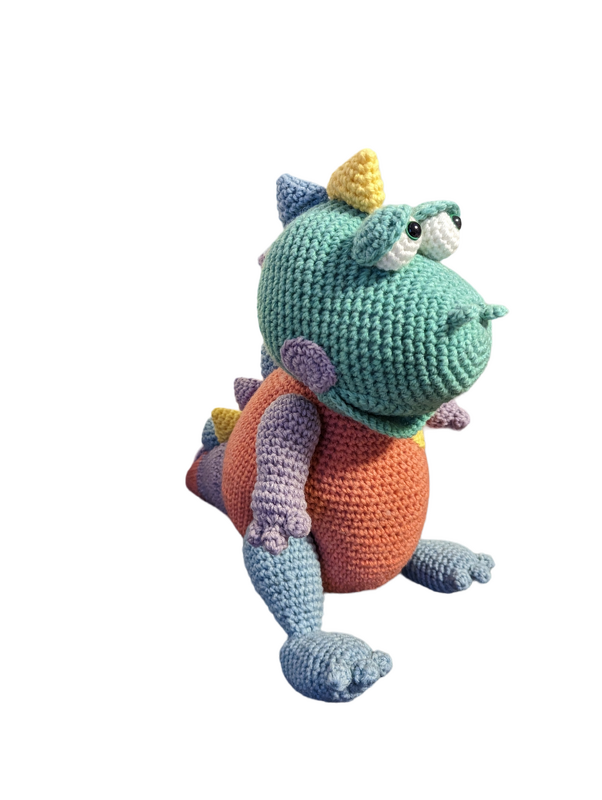 Handgefertigter sitzender Amigurumi-Dino mit türkisfarbenem Kopf, orangefarbenem Bauch und hellblauen Beinen auf neutralem Hintergrund.