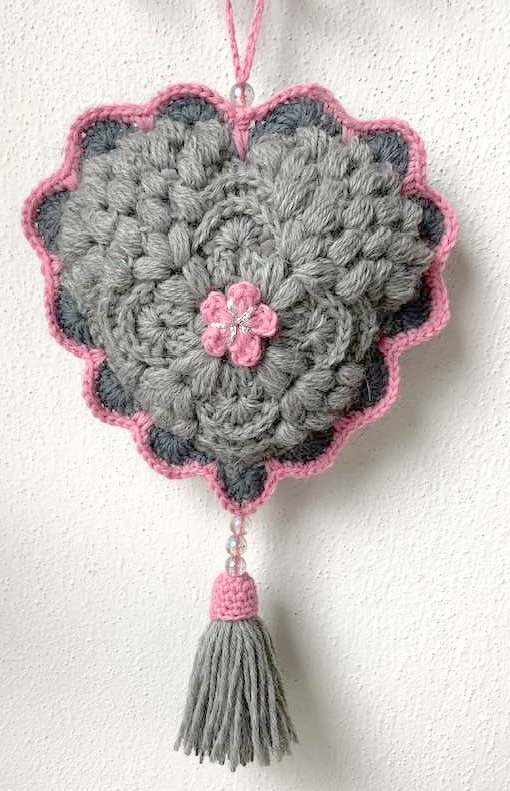 Gehäkelter Blumenanhänger in Grau und Rosa mit zentraler Blüte, Perlenakzent und rosa Quaste