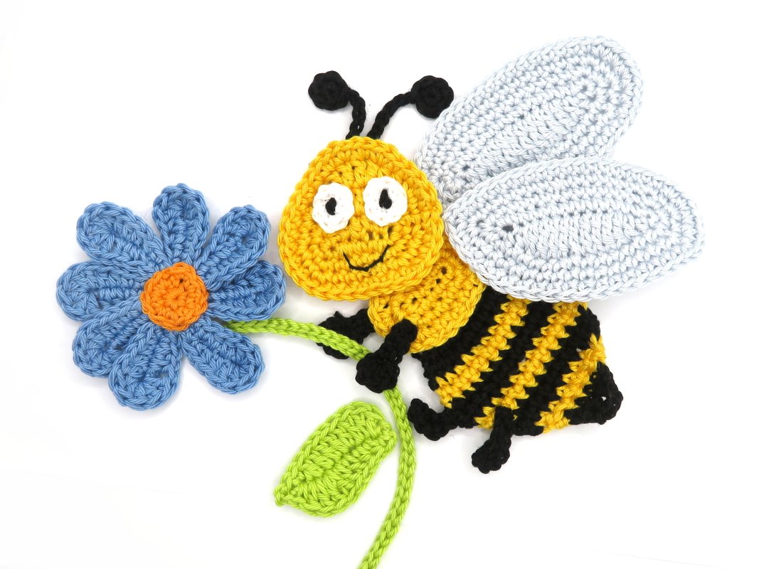Flat crochet bee applique in yellow and black holding a blue crochet flower on white background