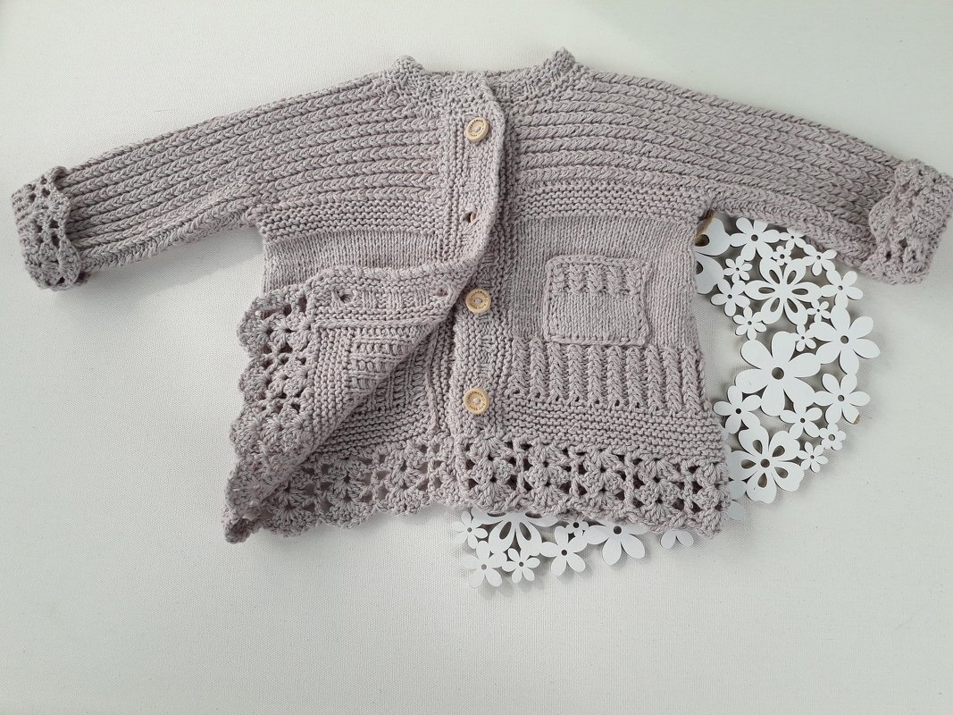 Hellgraues gestricktes Babyjäckchen mit runden Knöpfen, Rippen- und Lochmustern, aufgesetzter Tasche und gehäkelter Spitzenkante auf hellem Hintergrund mit Papierblumen