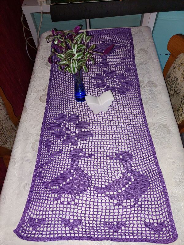 Violetter gehäkelter Tischläufer im Filetmuster auf hellem Tischtuch, mit blauer Vase und Zimmerpflanze