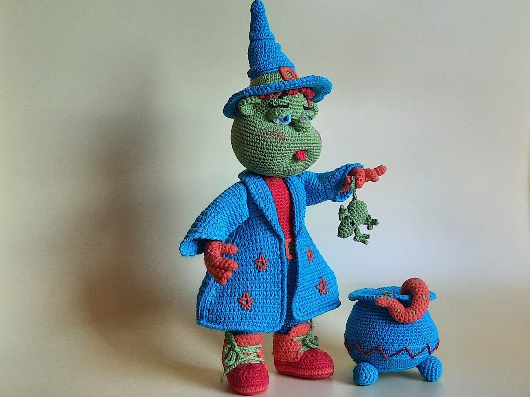 Gehäkelte grüne Hexe mit blauem Hut und Mantel hält einen Frosch, daneben ein blauer Kessel.
