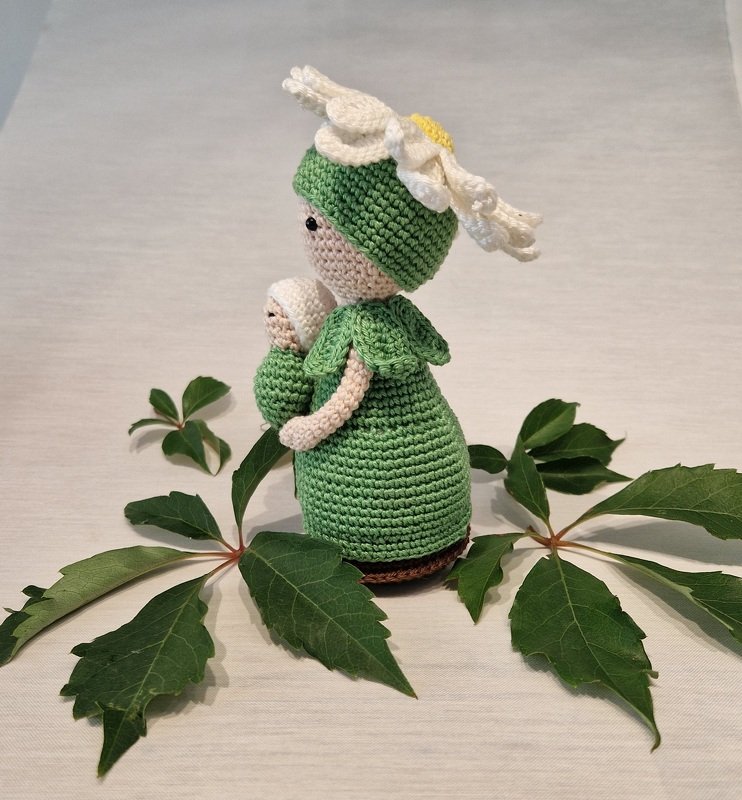 Handgehäkelte Puppe in Grün mit weißem Gänseblümchen auf dem Kopf, die eine kleine gehäkelte Baby-Puppe hält, arrangiert mit grünen Blättern.