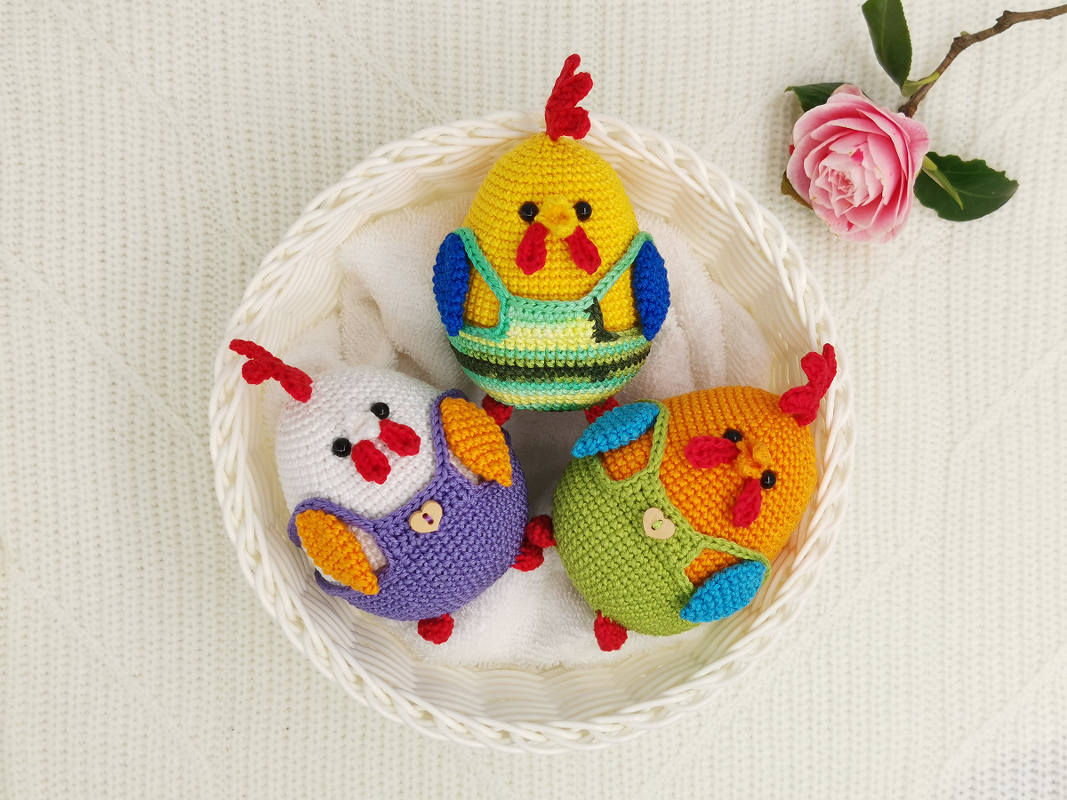 Three colorful crocheted chick amigurumi toys in a white basket with a pink rose on a knitted white blanket