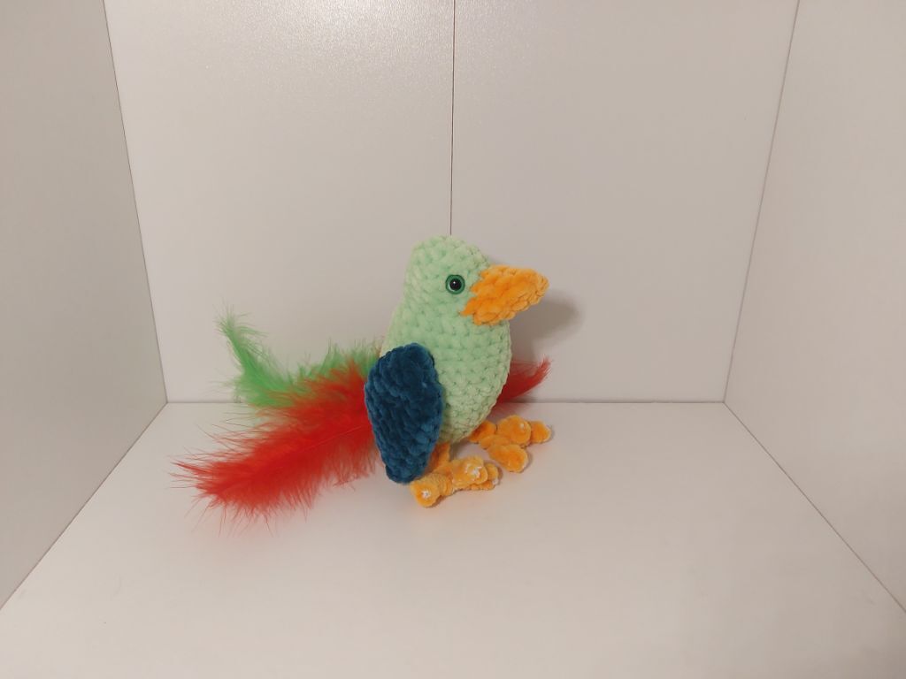 Gehäkelter hellgrüner Vogel mit orangefarbenem Schnabel, blauen Flügeln und buntem Feder-Schwanz auf weißem Hintergrund