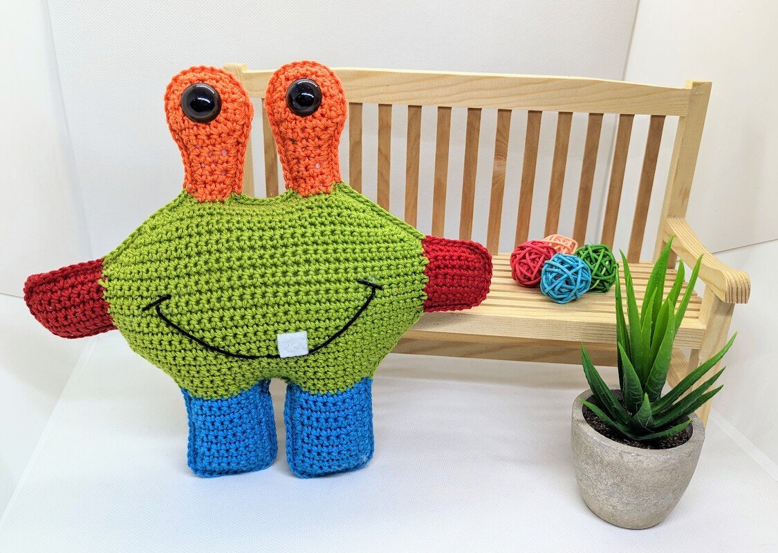 Buntes gehäkeltes Amigurumi-Monster mit Stielaugen vor kleiner Holzbank und Topfpflanze.