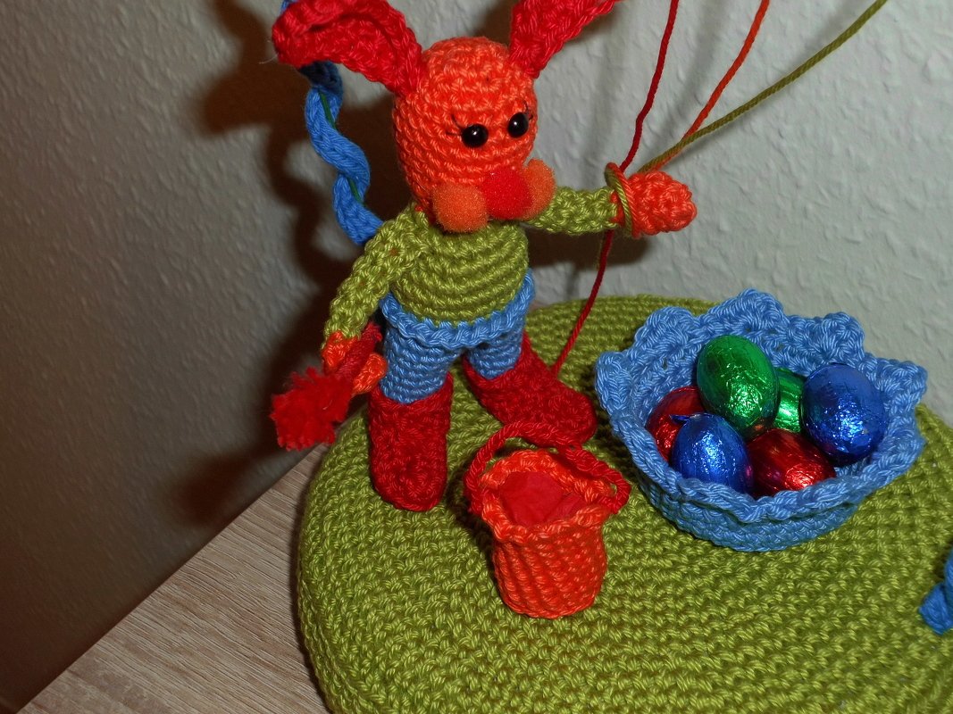 Gehäkelter oranger Hase mit grüner Oberbekleidung, blauen Hosen und roten Stiefeln, dazu ein kleiner roter Eimer und ein blaues Körbchen mit bunten Folieneiern auf grün gehäkelter Unterlage.