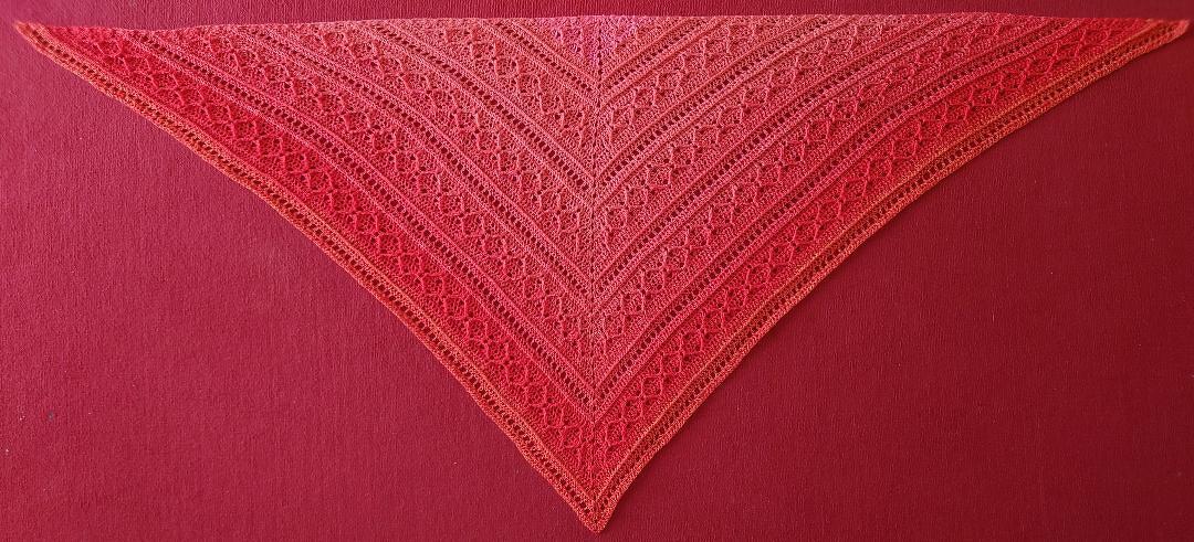 Rotes bis orangefarbenes gehäkeltes Dreieckstuch mit Lochmuster, flach auf rotem Hintergrund gelegt.